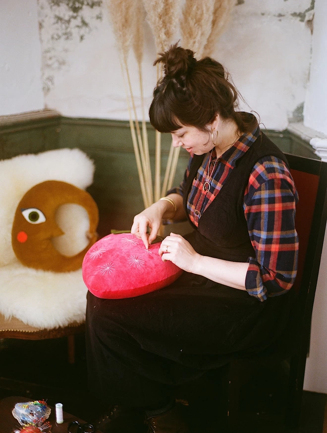 Woman with dark hair sits and sews a pink cushion 