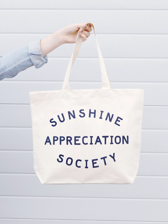 arm holding out a large natural canvas tote bag bearing the words sunshine appreciation society 