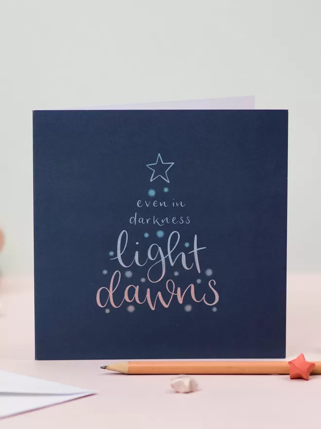 A square dark blue card stands on a pastel backdrop. The card features a lettered design which reads: 'Even in darkness, light dawns' in the shape of a Christmas tree