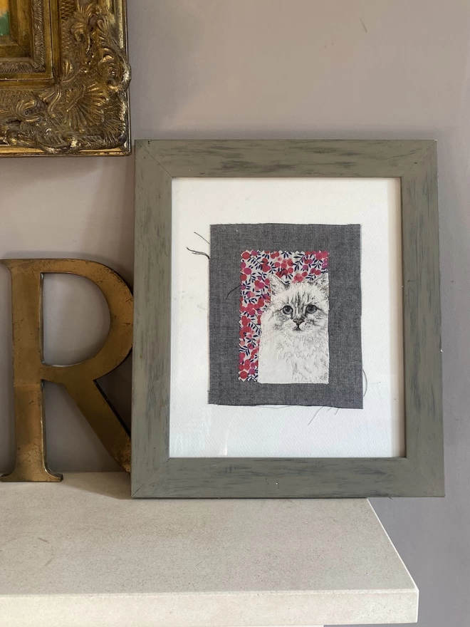 embroidered pet portrait of a white cat, framed and on mantlepiece
