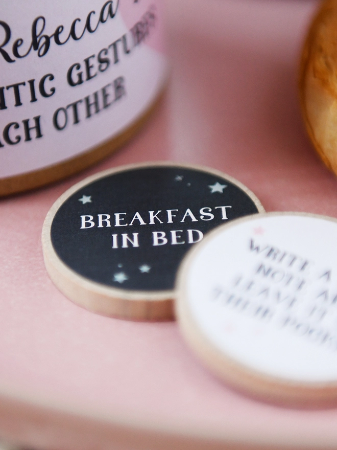 Two wooden tokens rest on a pink surface, the main one has a grey label and the words "Breakfast in bed" printed on it.