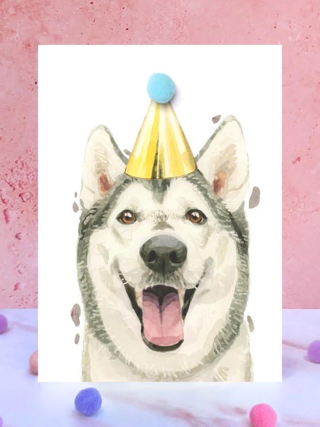 A greeting card featuring a hand painted design of a siberian husky, stood upright on a marble surface surrounded by pompoms. 