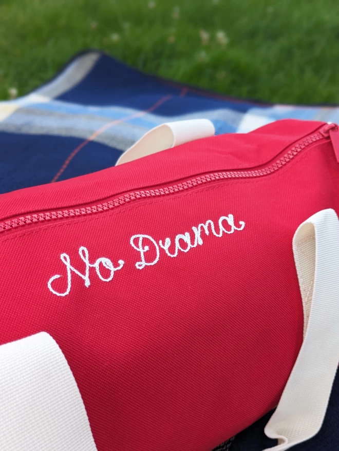 Red Duffel Bag with white straps and handles, placed on blue checkered picnic blanket in a field on a summers day, with a green grass background. The bag features white embroidered cursive lettering reading 'No Drama'