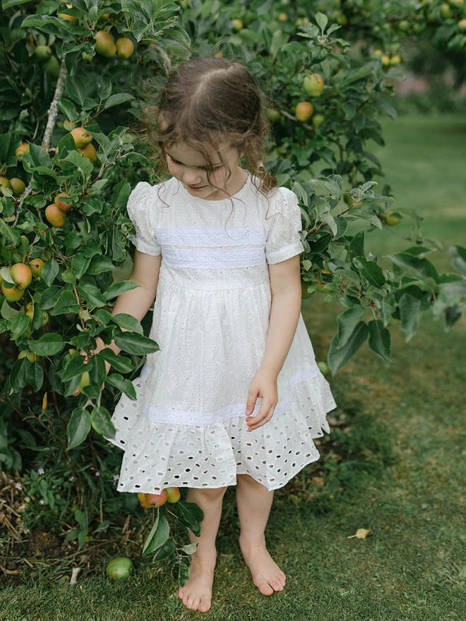 A little girl in a dress of contrasting white broderie anglaise fabrics stands beside a tree