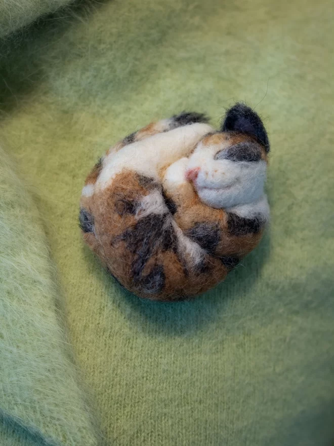 A needle-felted sleeping kalico cat brooch on a green fluffy jumper