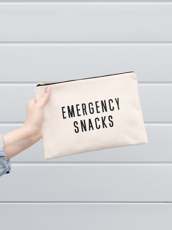a hand holding out a natural canvas zip pouch with the words emergency snacks
