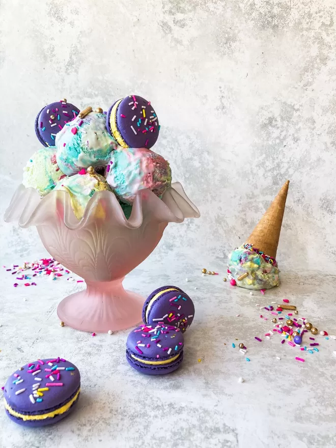 a pink glass bowl filled with colourful ice cream balls, topped with bright purple macarons and colourful sprinkles