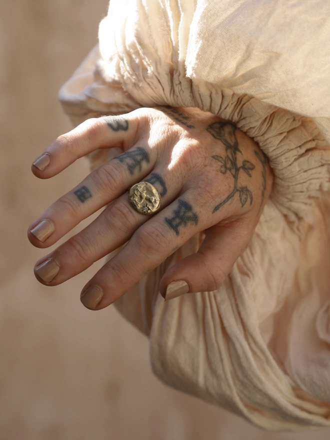 Image of tattooed female hand wearing a puffed linen fabric sleeve and a brass ring with the design of a horse on it. The background of the image is a terracotta rough plaster 