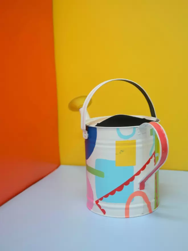 Hand painted watering can by Julie-Anne Pugh. Base colour is white with bring coloured shapes weaving across the body of the can on a yellow and orange background. Photographed from the back.
