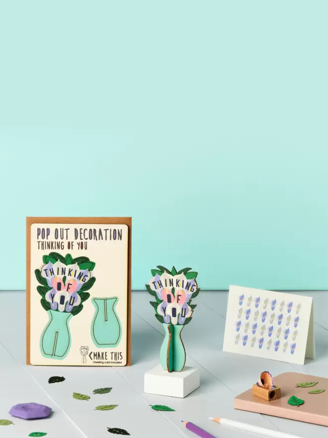 3D laser-cut thinking of you pop out decoration and lily pattern card and brown kraft envelope on top of a wooden desk in front of a pale blue coloured background