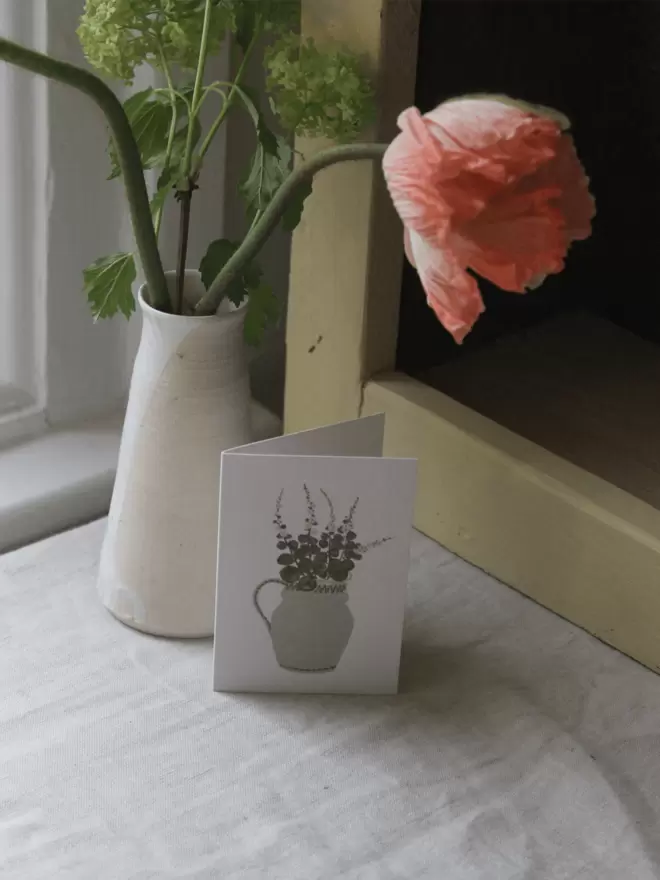 mini greetings card with cut hollyhocks on it arranged in a light green vase, next to a vase displaying Icelandic poppies. 
