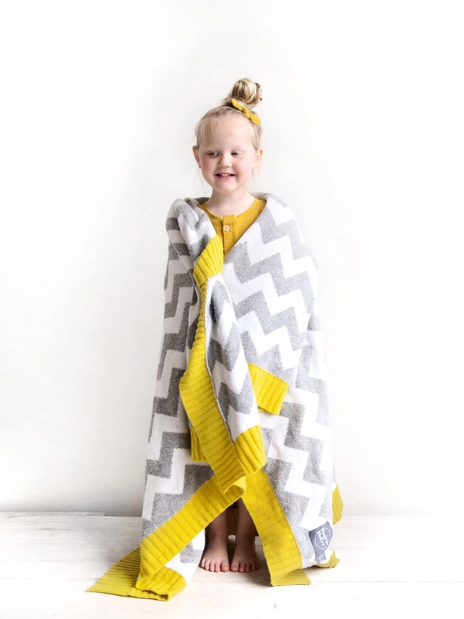 A little girl stand against a white background, draped in a grey and white knitted blanket with a chevron design and a yellow trim. She is smiling and has a top knot in her hair with a matching yellow scrunchie around it.
