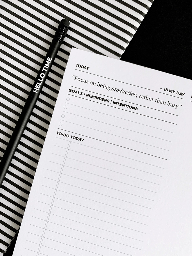 A close up of a My Day Daily Productivity Pad laid next to a HELLO TIME pencil on a black and white striped background.