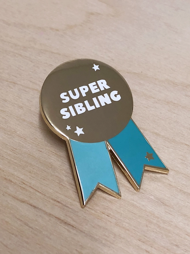 A turquoise and gold pin badge in the shape of a rosette is on a wooden table. It has the words "Super sibling" on.