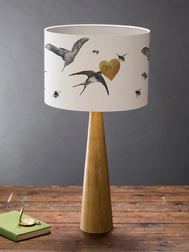 Drum Lampshade featuring birds and bees with a gold heart on a wooden base on a shelf with books and ornaments