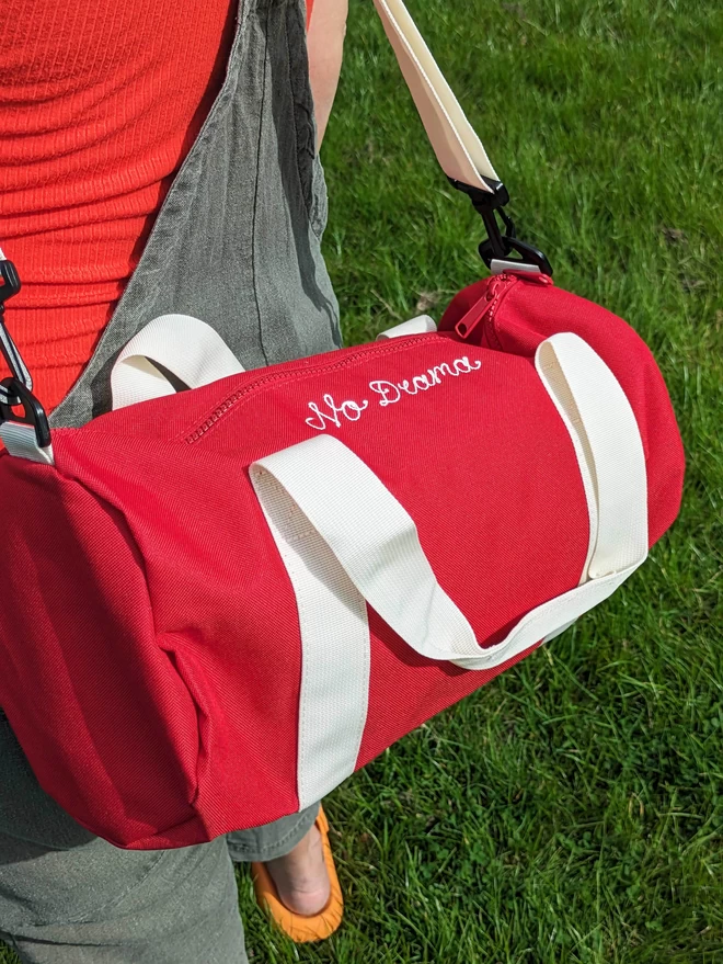 Red Duffel Bag with white straps and handles, being worn by a woman on a green grass background. The bag features white embroidered cursive lettering reading 'No Drama'