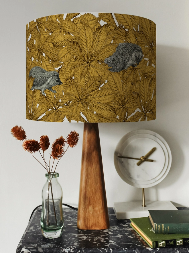 Drum Lampshade featuring hedgehogs in yellow autumn leaves on a wooden base on a shelf with books and ornaments