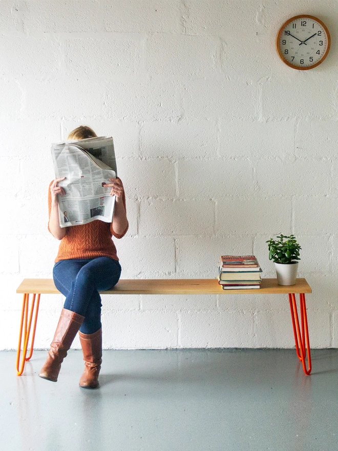 woman sat reading a newspaper on a hairpin leg bench with oak seat and orange hairpin legs