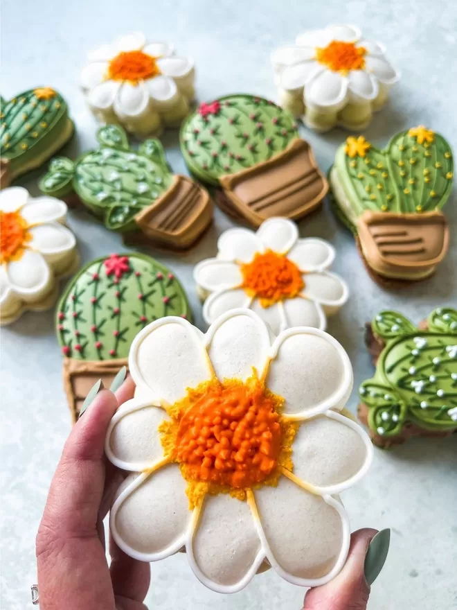 Large cactus shaped macarons decorated with yellow, pink & white flowers and white Daisy shaped macarons on a cement background