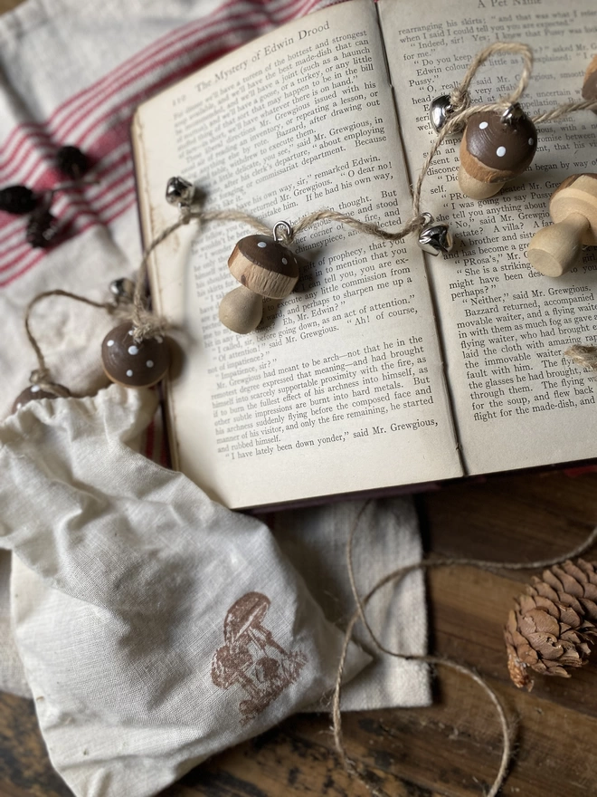 A string of Hand Painted Wooden Toadstool Bell Garlands trailing across an opened book atop a red and white striped cloth, on display with a small white pouch and some decorative pine cones