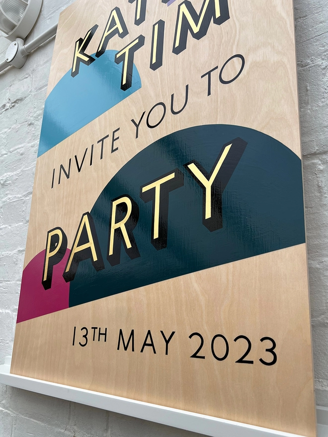 Handpainted invite to party sign with gold leaf letters outlined in black, with bright coloured semicircles in the background, against a white brick wall. 