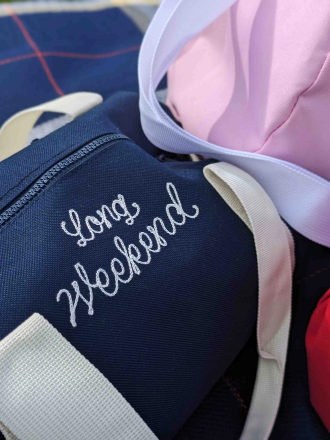 Navy Blue Duffel Bag with embroidered 'Long Weekend' sitting on a blue checkered picnic blanket in a field on a summers day