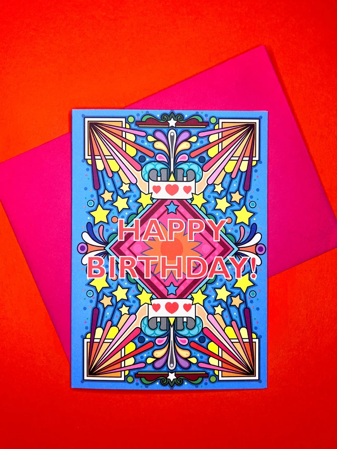 A vibrant blue birthday card with a bold multi-coloured pattern, with Happy Birthday at the centre, sits on top of a pink envelope on a red backdrop.