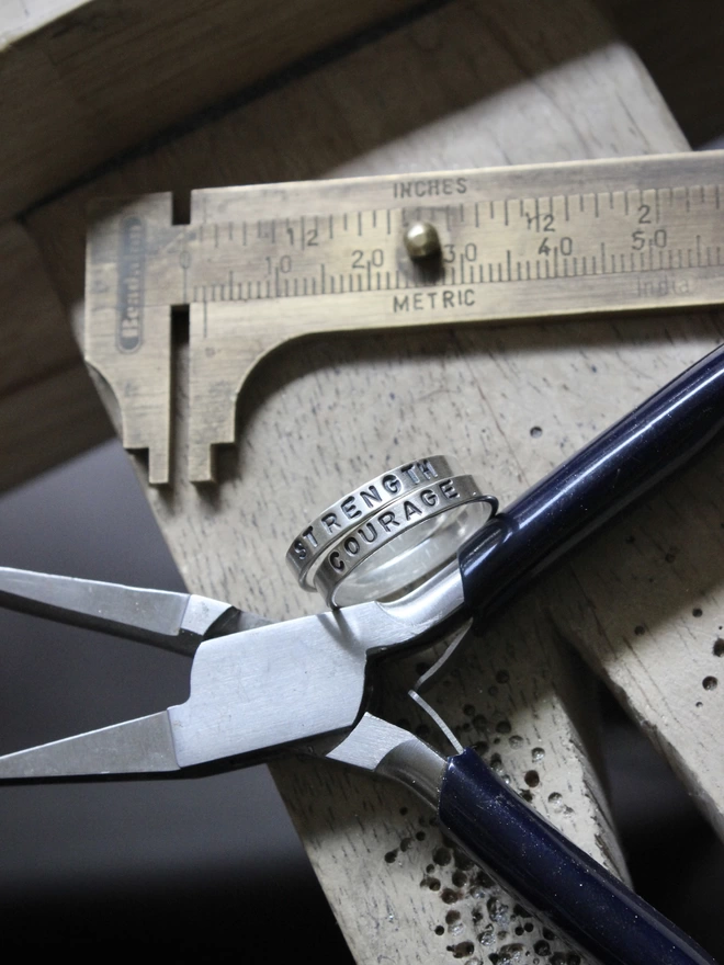 A pair of simple silver plain band rings with the words 'strength' and 'courage' stamped on them, resting against a pair of jewellery pliers on a jewellers wooden bench peg, with brass callipers lying behind.