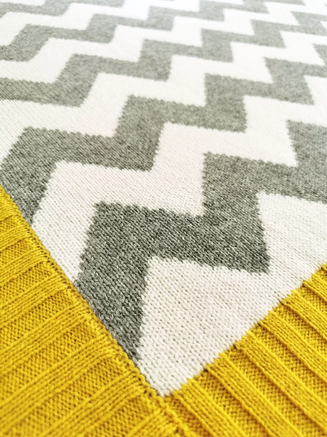 A close up of the corner of a knitted blanket showing the knit stitches, grey and white zigzag design and mustard yellow ribbed trim.