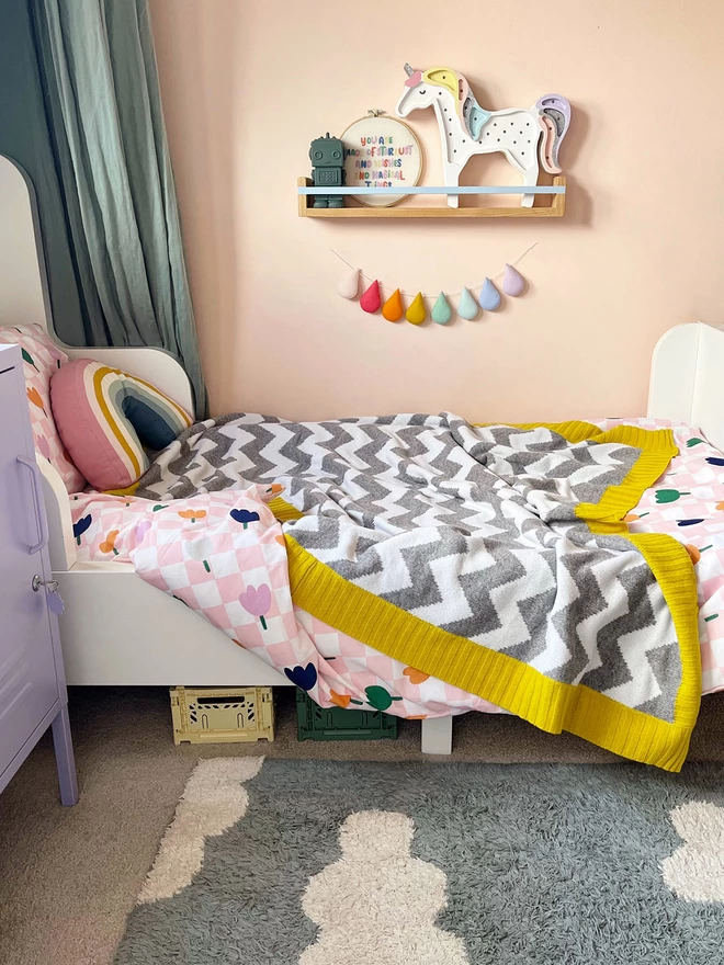 A view of a bright and colourful childrens bedroom in muted rainbow colours showing a single bed with a grey and white chevron blanket with mustard yellow trim draped and ruffled over the bed.
