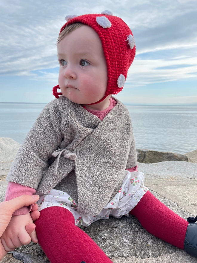 A baby in a knit and red tights wears a red crocheted bonnet with white dots