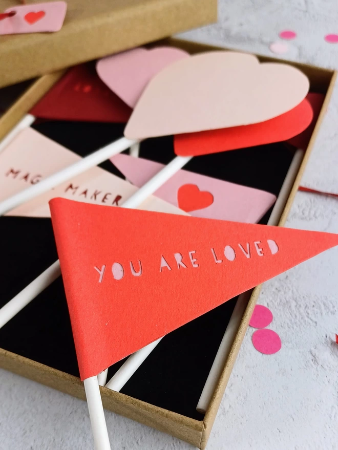 A closeup of a red triangular flag papercut to reveal the words "you are loved" in pink