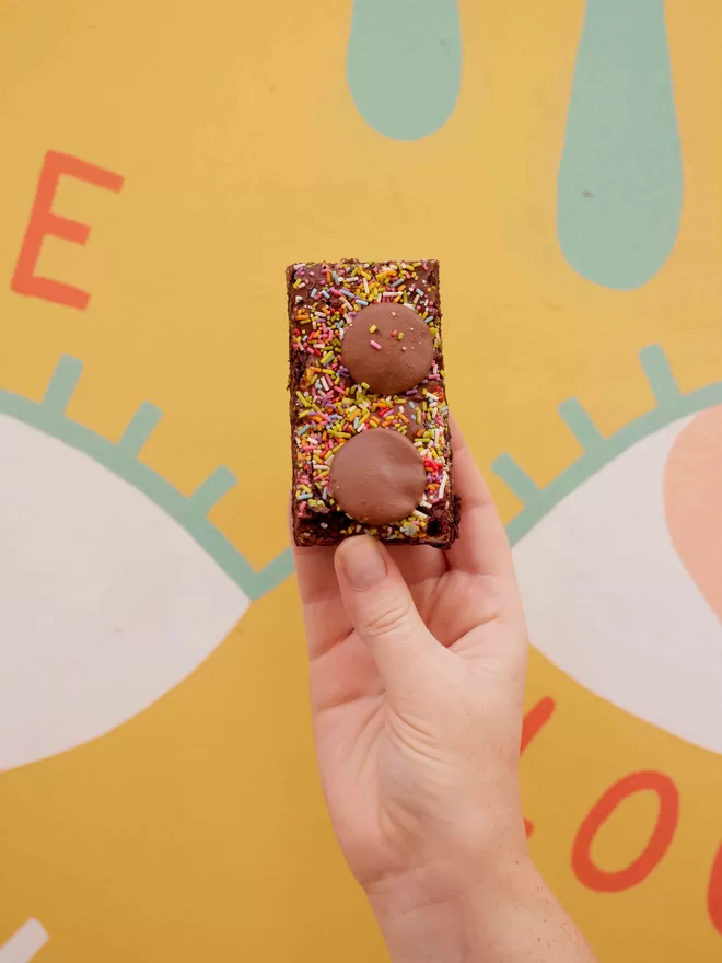 Hand holding a single Funfetti brownie slice with sprinkles and milk chocolate buttons set against a colourful background
