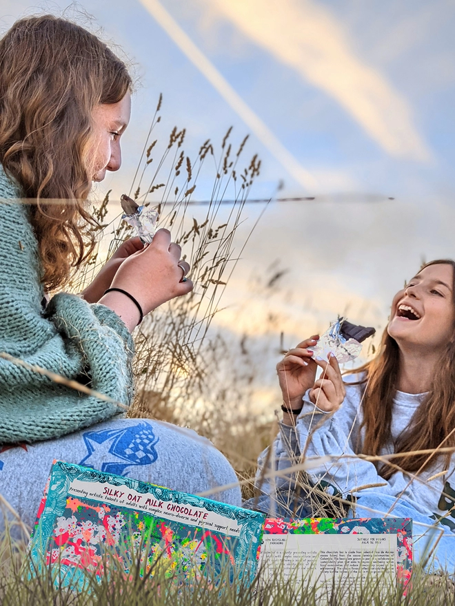 Two happy girls in a field enjoying charity vegan chocolate bar wrapped in silver foil 