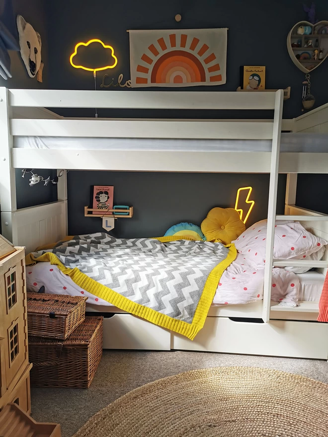A view of a moderns childrens room showing white bunk beds against dark grey walls. On the lower bunk bed is a grey and white zigzag blanket with a yellow border. Lightning bolt and cloud neon nighlights glow yellow against the dark walls creating a cosy vibe.