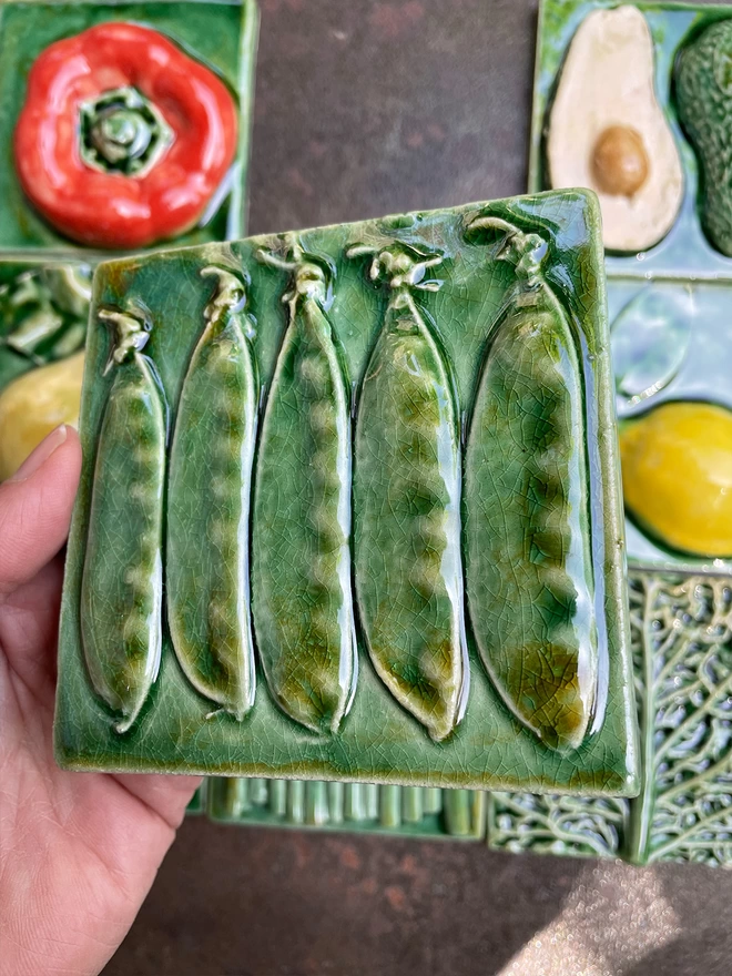 Mange tout tile - square, 3D, realistic and glossy. Five mange touts are lined up on a lush green glaze background. Other fruit and vegetable tiles in the series are on display in the background: lemon, fig, savoy cabbage, pear, red capsicum pepper, avocado, garlic, asparagus.
