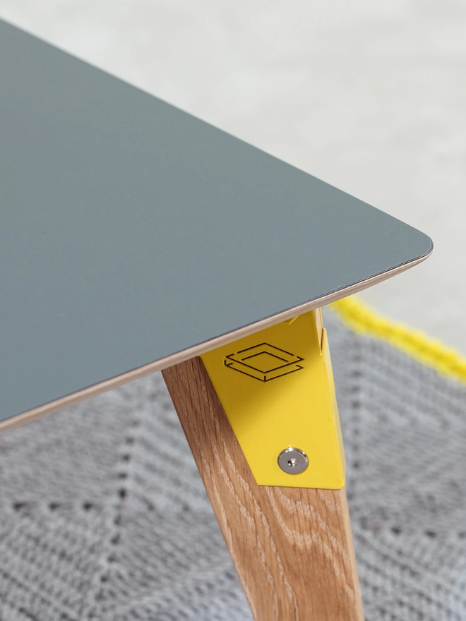Corner view of a stylish and minimalist coffee table with grey Fenix top, yellow coloured steel brackets and solid oak legs on a grey rug