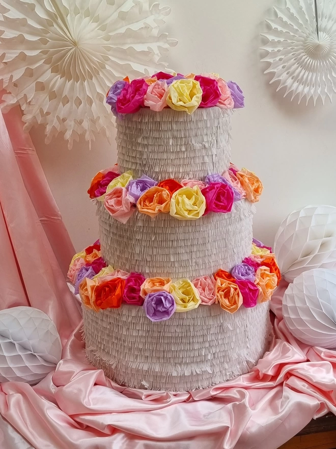 3 tier wedding cake pinata with colourful flowers topping each tier on a pink satin backdrop surrounded by white paper decorations