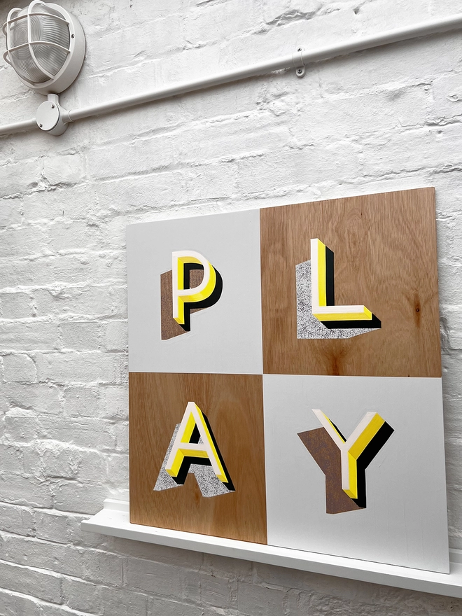 PLAY hand painted sign in neon yellow, lilac and grey, against a white brick wall, at an angle. 