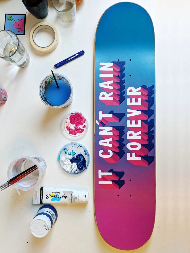 A skateboard that has a spray painted pink and blue background and 3d typography that reads It Can’t Rain Forever in pink and white lies on a desk alongside paint tubes, palettes, paintbrushes and tools. Artwork by Survival Techniques