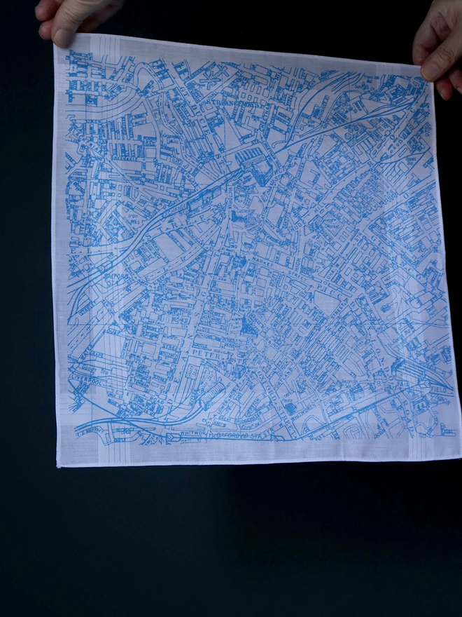 A Mr.PS Manchester map hankie printed in sky blue held aloft by two hands in front of a dark wall 