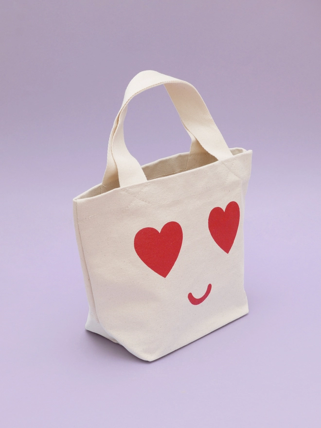 a mini kid's size tote bag with heart eyes and smiling mouth standing on a purple backdrop