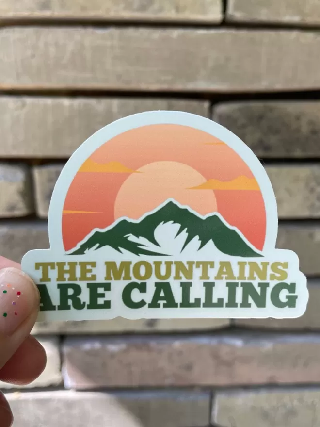 The Mountains Are Calling Vinyl Sticker being held in front of a wall.