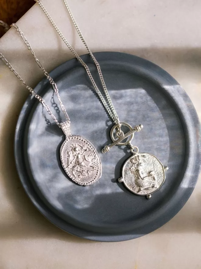 Silver oval neckclace next to silver coin pendant with toggle featuring goddesses on blue background