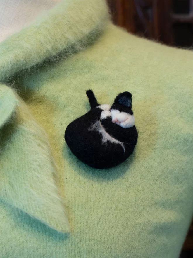A needle feltedblack and white sleeping cat brooch on a green fluffy jumper 