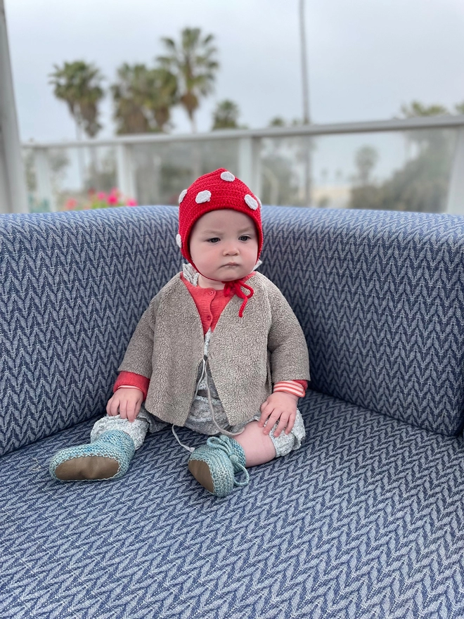 A baby in a knit wears a red crocheted bonnet with white dots