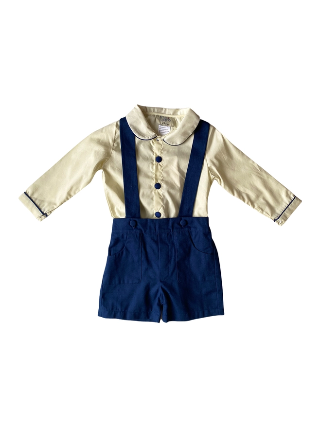 Cutout of a cream shirt with a peter pan collar and navy piping and buttons and navy shorts with braces