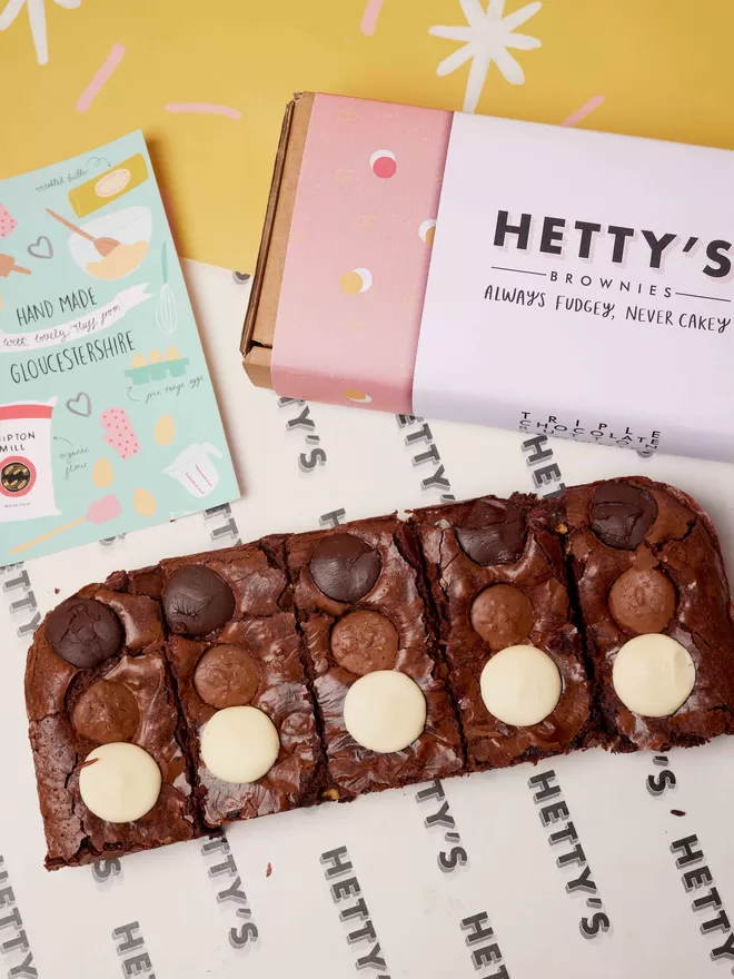 Five slices of triple chocolate brownies in a flat lay with branded box and postcard