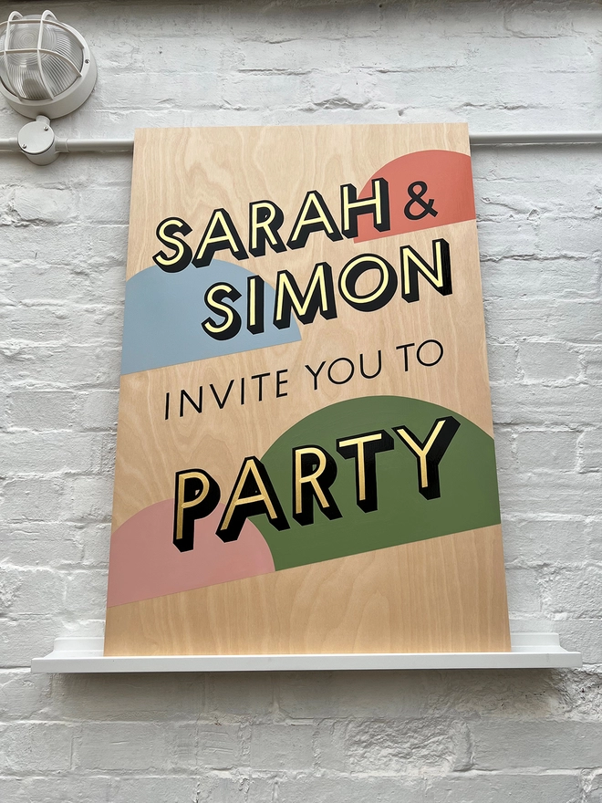 Handpainted invite to party sign with gold leaf letters outlined in black, with coloured semicircles in the background, against a white brick wall. 