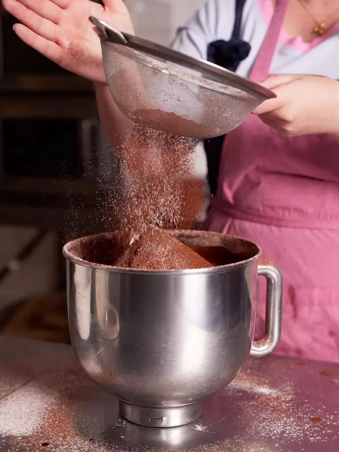 Founder sieving to create the brownie base mixture in the bakery wearing a pink apron